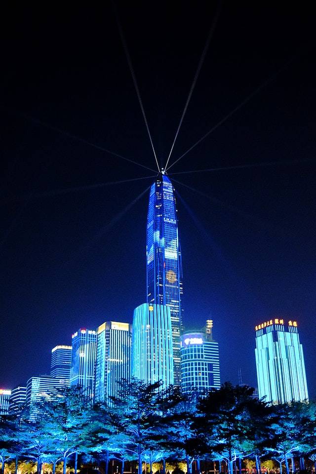 A skyscraper at night illuminated with blue lights and beams radiating from its spire against a dark sky.